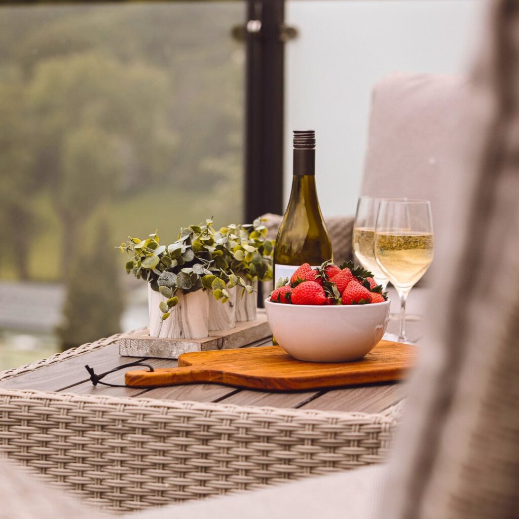 A bottle of champagne, two champagne flutes with champagne in and a bowl of strawberries on a table top. The shot is a close up of these elements on the decking of one of the caravans.