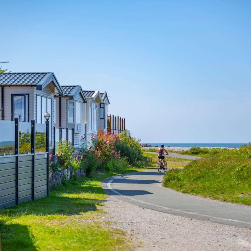 Big blue sky, a row of caravans on the left hand side with a path to their right. There is a woman on a bike cycling away from the camera and to the right of her in the distance is the sea.