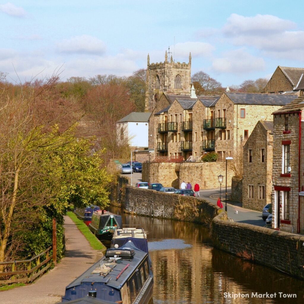A picture of Skipton Market Town. On the left there is a canal-side footpath leading away from the viewer, down the centre of the image is the canal with a canal boat moored in the forefront of the image. To the right of the canal is the town, with a few stone cottages and a church.