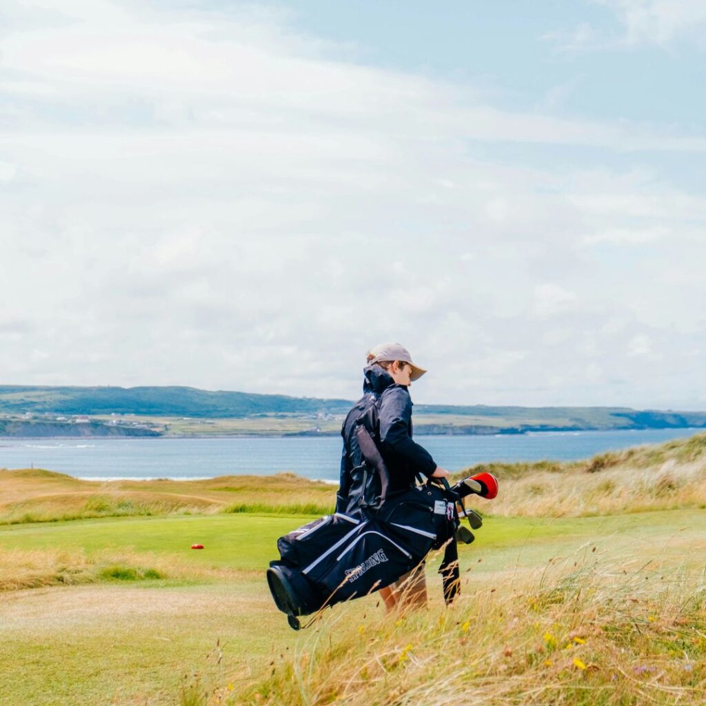 Golfer walking across a coastal links golf course with sea views.
