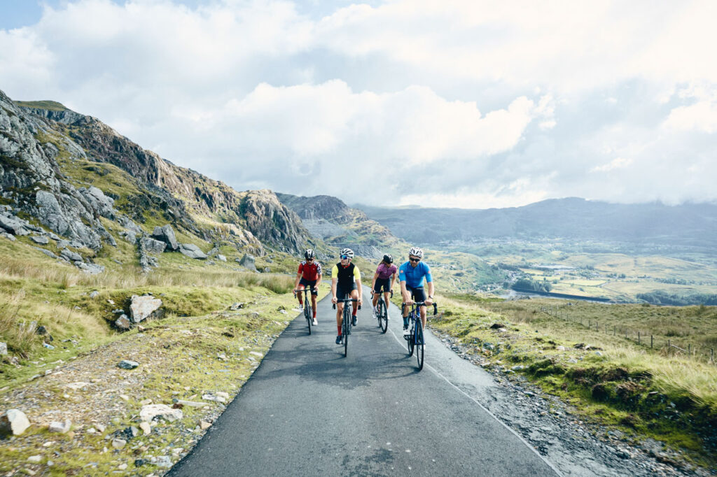 Four road cyclists climbing a mountain road beside rocky slopes, with a valley below.