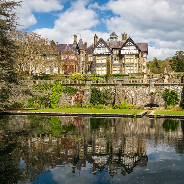 Bodnant Hall reflected in a garden pool, with stone terraces, trees and blue sky.