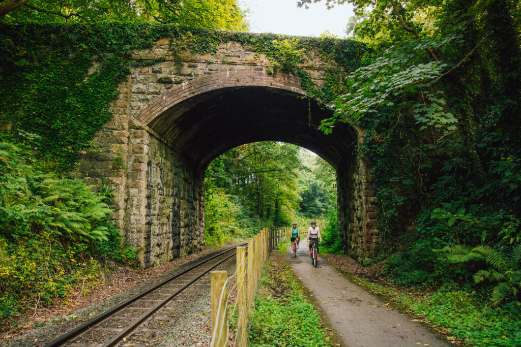 Two cyclists following a wooded path beside a railway line beneath a stone arch bridge.