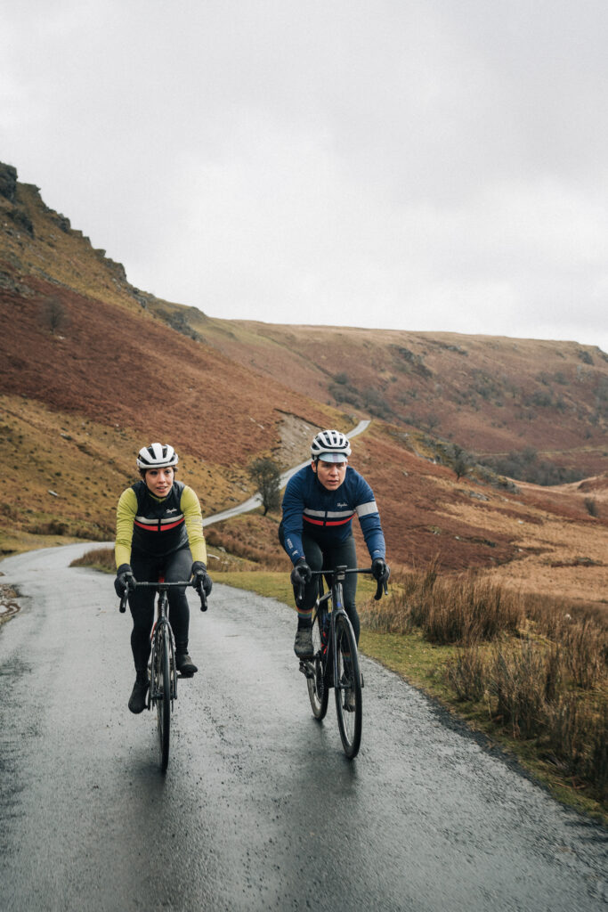 Two road cyclists climbing a wet country road beneath rugged hills.