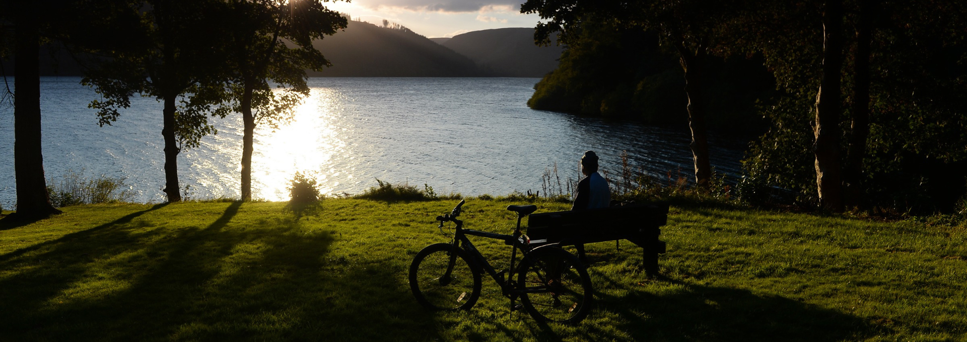 Cyclist resting on a bench beside a lake, with a bicycle in the foreground and sunlight reflecting on the water.